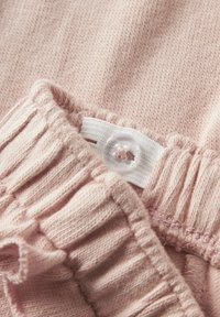 Tissu tricoté rose pâle avec un bord froncé et une taille élastique, comportant une étiquette blanche maintenue par un bouton transparent. Texture douce.