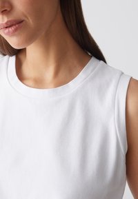 Nahaufnahme einer Frau, die ein ärmelloses weißes Shirt mit Rundhalsausschnitt trägt, das ihren Hals, ihre Lippen sowie einen Teil ihrer Schulter und Haare zeigt.