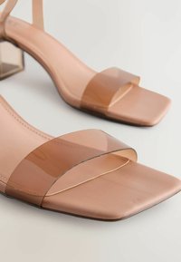 Läpinäkyvät korkokengät neliönmuotoisella kärjellä. Valmistettu muovista ja synteettisestä materiaalista, nude-värinen ja minimalistinen muotoilu.