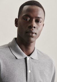 Graues Poloshirt mit Kragen, aus strukturiertem Stoff und drei weißen Knöpfen, vor einem neutralen Hintergrund präsentiert.