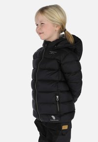 Young child with blonde hair in a ponytail wearing a black puffer jacket with a hood, standing sideways, hands behind back, smiling slightly.