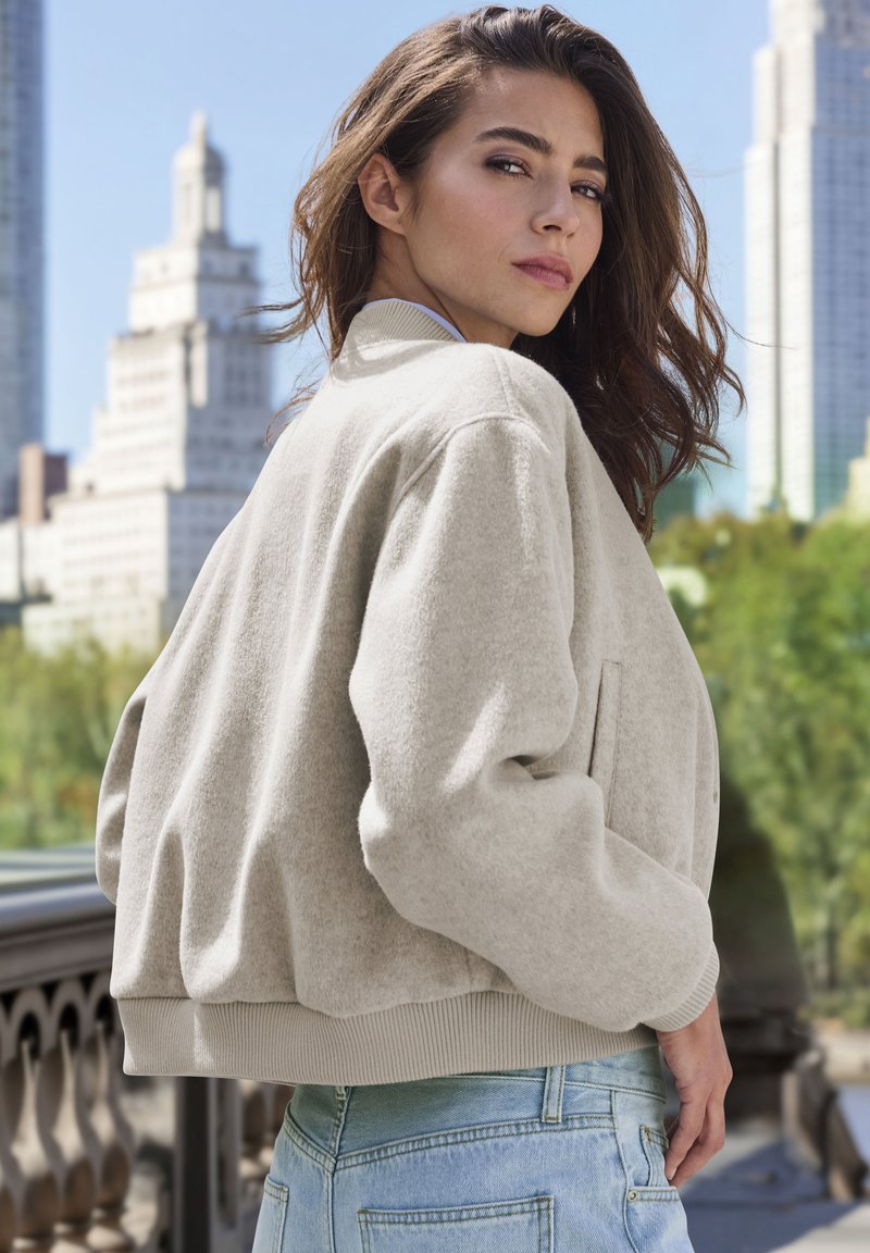Light gray fleece bomber jacket with ribbed cuffs and waistband, featuring side pockets and a relaxed fit. Worn over light blue denim.