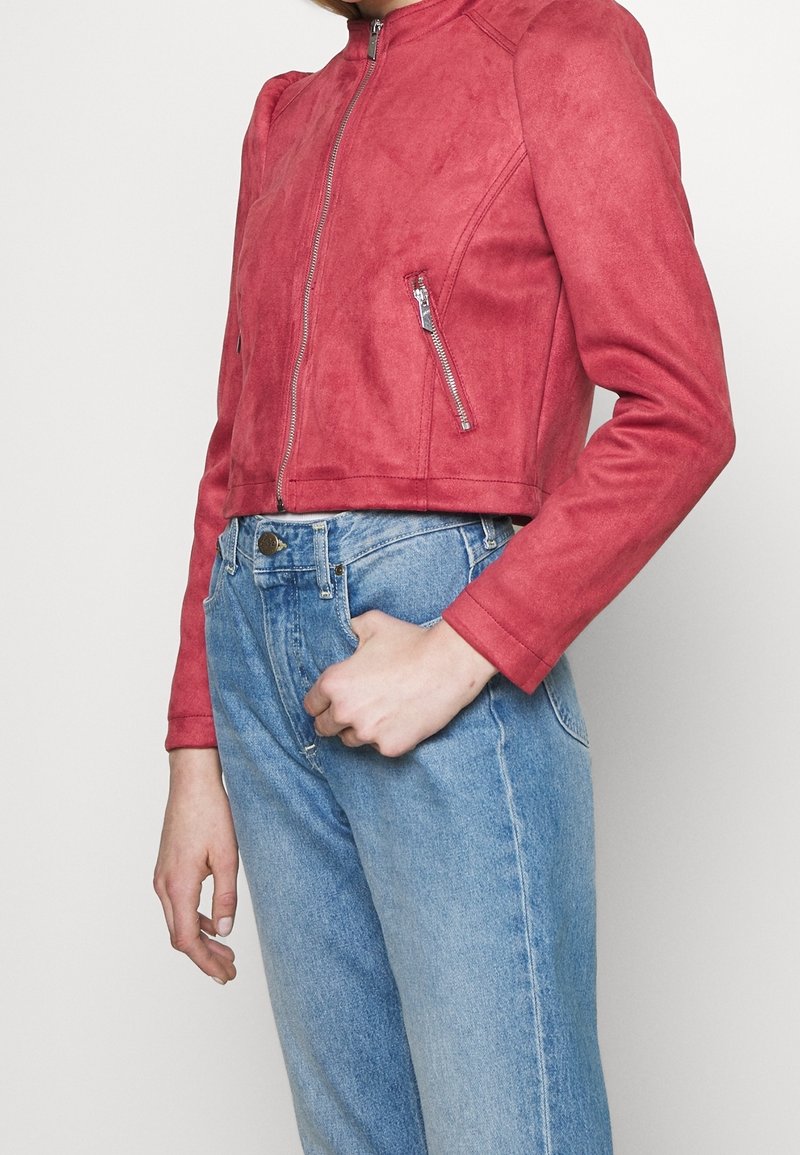 Personne portant une veste courte zippée en daim rouge et un jean taille haute bleu clair, avec une main reposant près de la poche.