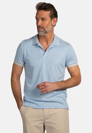 Man met grijze baard, die een lichtblauw poloshirt met korte mouwen draagt met gestreepte mouwboorden en beige broek, staand met één hand in de zak.