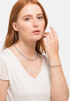 Collana di metallo argentato attorcigliata e bracciale coordinato su una donna di carnagione chiara. Indossa un top bianco a scollo a V. Texture liscia, design casual.