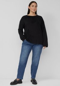 Frau mit langen dunklen Haaren, die einen schwarzen Pullover mit Blumenmuster, blaue Jeans und schwarze Loafers trägt, steht vor einem einfarbigen hellen Hintergrund.