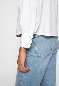 Personne portant un jean bleu clair et une chemise blanche avec des poignets en feston et des boutons en perles, la main détendue sur le côté.