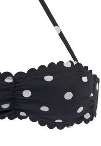 Zwart bikinitopje met witte stippen, met gekartelde randen en dunne bandjes. De stof is rekbaar en textuur heeft. Gedetailleerde ruching is zichtbaar.