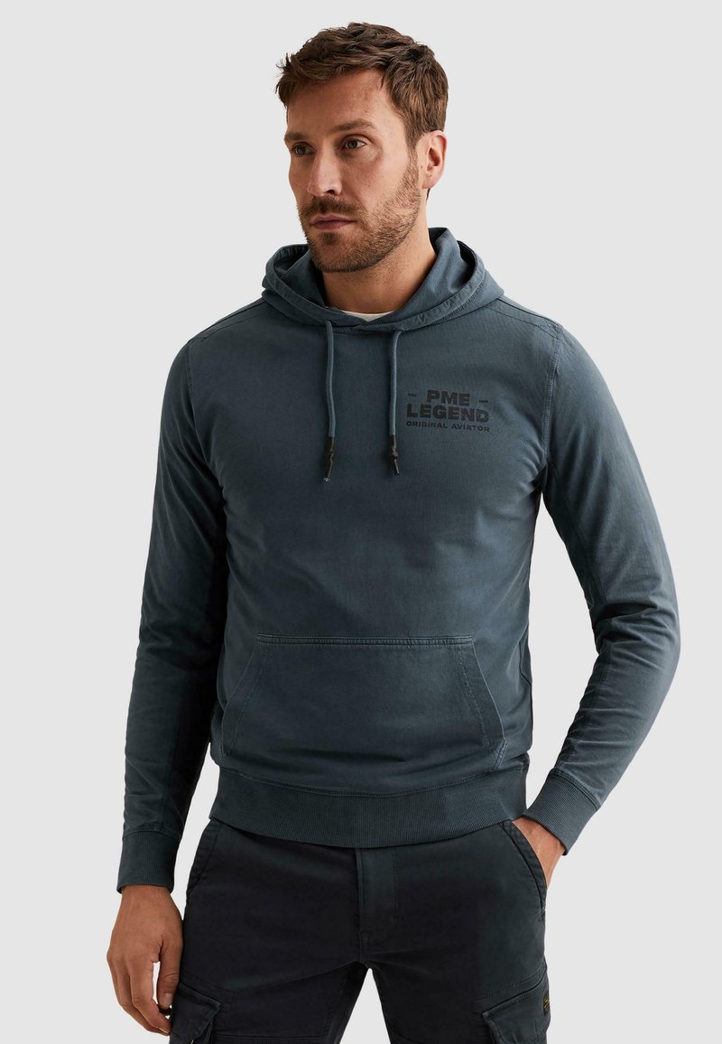 Homme portant un sweat à capuche bleu foncé avec poche avant et logo PME Legend, posant avec une main dans la poche devant un fond uni.