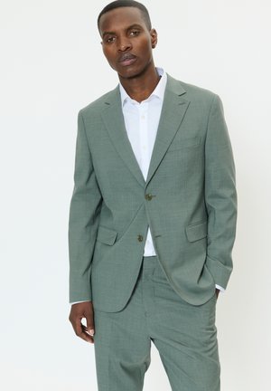 Jasnozielony garnitur z teksturowanej tkaniny. Zapięcie na jeden guzik, klapy z wycięciami i boczne kieszenie. Noszony z białą koszulą pod spodem.