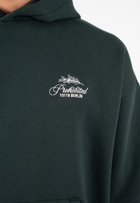 Felpa verde scuro con testo ricamato bianco "Proibito 10119 Berlino" e un piccolo disegno di foglia sopra il testo.