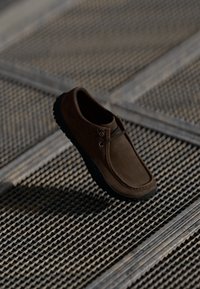 Scarpa in suede marrone con punta arrotondata, suola in gomma nera e due occhielli per il lacci, posizionata sopra una superficie grigliata texturizzata.