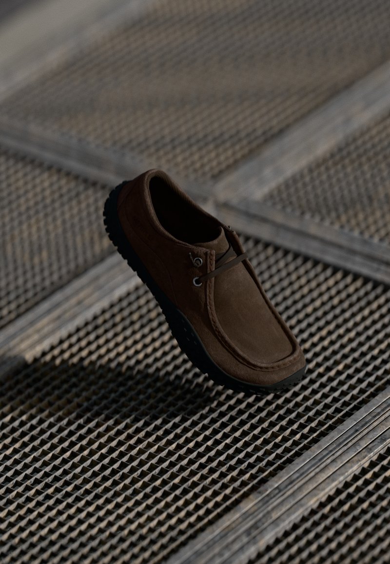 Scarpa in suede marrone con punta arrotondata, suola in gomma nera e due occhielli per il lacci, posizionata sopra una superficie grigliata texturizzata.