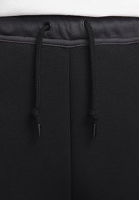 Schwarze Sweatpants aus einem strukturierten Stoff mit einem Kordelzugbund, akzentuiert durch schwarze Metallteile. Der Bund hat eine glatte Oberfläche.