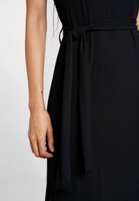 Czarna prążkowana sukienka z wiązanym paskiem w talii, prezentująca gładką teksturę. Widać rękę z minimalistycznym tatuażem na nadgarstku.