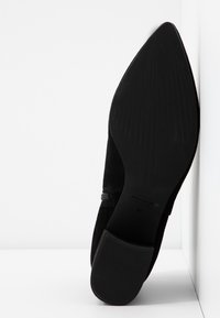 Chaussure en daim noir avec un bout pointu et un design minimaliste. Elle présente une semelle en caoutchouc lisse et plate avec des rainures subtiles pour l'adhérence.