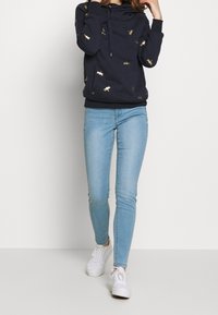 Felpa blu marina con motivi d'insetti dorati, collo con cordini e orlo a coste, abbinata a jeans skinny blu chiaro e sneakers bianche.