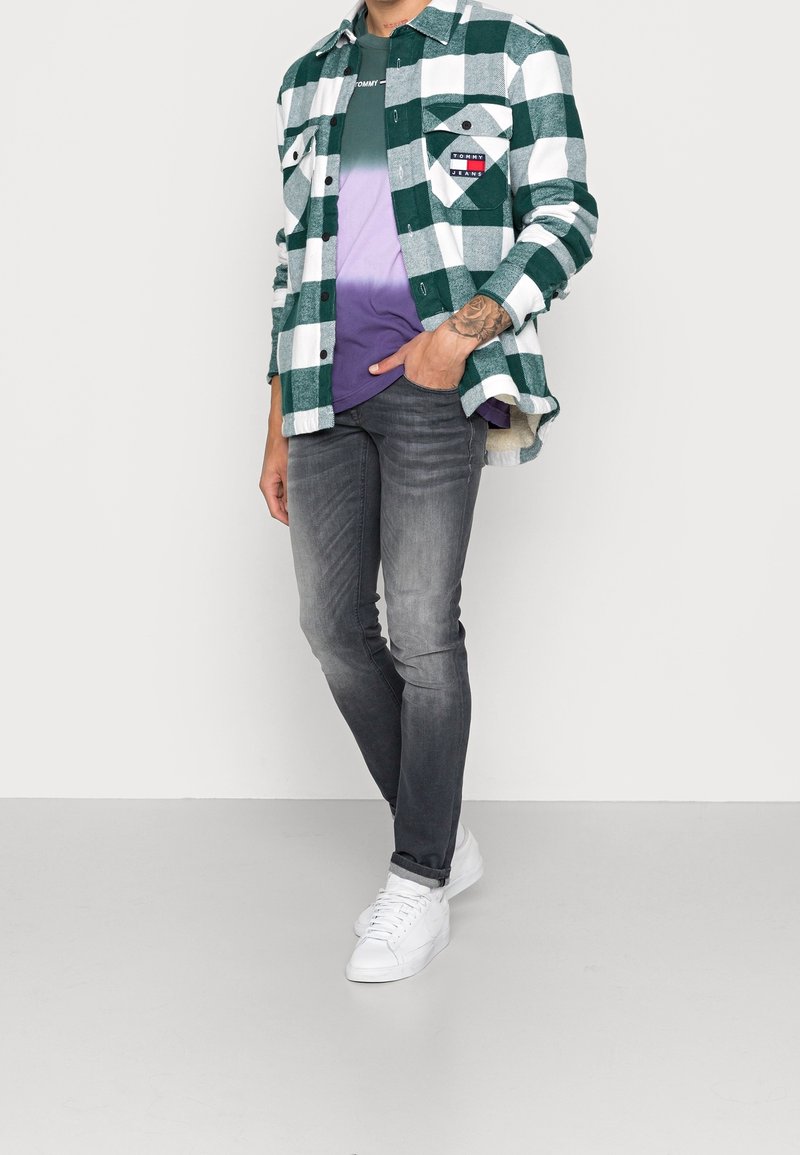 Homme portant une chemise à carreaux verte et blanche sur un t-shirt dégradé violet, jean noir délavé et baskets blanches, main dans la poche.