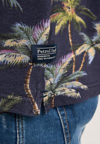 Camisa de manga corta con una tela azul marino y un estampado colorido de palmeras. Incluye una etiqueta azul cosida en el dobladillo.