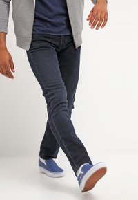 Scarpe slip-on di tela blu, jeans di denim scuro a taglio slim, felpa grigia con zip sopra una maglietta blu navy; visibile movimento di camminata.