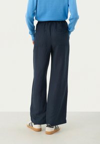 Pantaloni a gamba larga blu navy con vita elasticizzata, due tasche posteriori, abbinati a un maglione azzurro chiaro e sneakers bianche con dettagli neri.