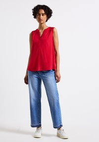 Top rosso senza maniche con scollatura a V e texture arricciata sulle spalle, abbinato a jeans a gamba larga blu chiaro e sneakers bianche.