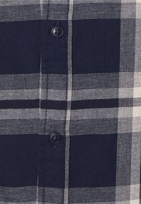 Camicia a quadri navy e bianca con chiusura a bottoni, caratterizzata da diverse dimensioni dei quadri e un tessuto texturizzato. Due bottoni scuri visibili sulla pattina.