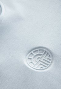 Helles blaues Gewebe mit glatter Textur, das ein geprägtes, kreisförmiges Logo mit geometrischen Mustern aufweist.