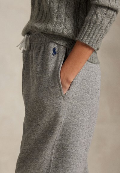 Person trägt grauen Strickpullover und graue Jogginghose, die Hand in der Tasche und ein sichtbares dunkelblaues Logo auf der Jogginghose.