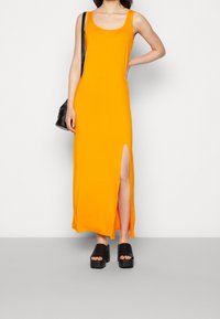 Pomarańczowa, bezrękawna maxi sukienka z bocznym rozcięciem, wykonana z gładkiego materiału. W połączeniu z czarnymi sandałami na platformie. Prosty, nieozdobiony design.