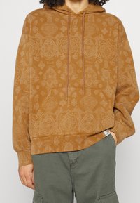 Persona viste una sudadera con capucha de color marrón con un patrón, que tiene un bolsillo frontal y cordones, combinada con pantalones cargo de color verde oliva, con la mano en el bolsillo.
