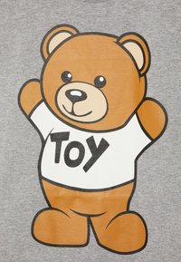 Hnědá medvědí hračka v bílém tričku s nápisem "Hračky", stojící na šedém pozadí, se vztyčenýma rukama a usměvavou tváří.