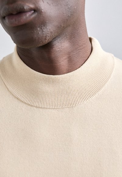 Beige gerippter Strickpullover mit hohem, rundem Kragen, der eine glatte Textur und eine enganliegende Passform am Hals präsentiert.