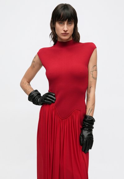 Czerwona sukienka z dopasowaną górą i rozkloszowanym, marszczonym dołem. Gładki, dzianinowy materiał z wysokim dekoltem, w połączeniu z czarnymi rękawiczkami.