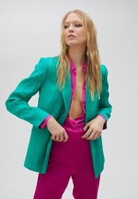 Blazer texturizado en turquesa con un escote en V profundo, combinado con una camisa de satén rosa brillante de botones y pantalones rosas a juego.
