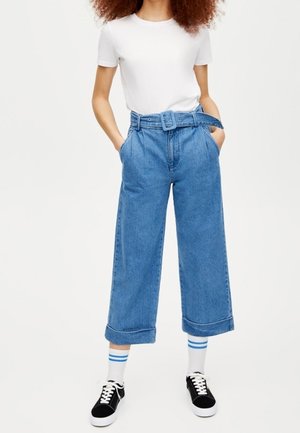 Persoon die een wit kortärmelig shirt draagt, een hooggetailleerde wijd uitlopende blauwe spijkerbroek met riem, witte sokken met blauwe strepen en zwarte sneakers.