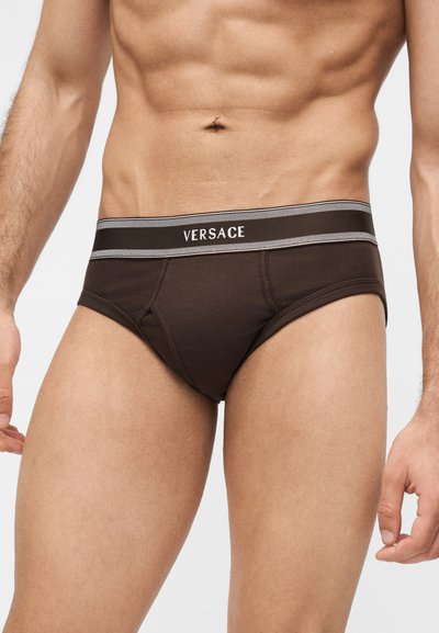 Torse masculin portant un slip Versace marron foncé avec une ceinture noire et grise, présentant un nombril percé, sur un fond clair uni.