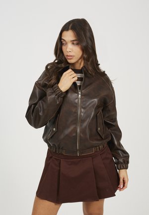 Giubbotto bomber in pelle marrone con zip frontale, tasche laterali, polsini ed orlo elasticizzati. Indossato sopra una maglietta a righe e abbinato a una gonna marrone.