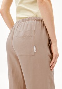 Beige Leinenhosen mit elastischem Bund, einer Gesäßtasche und einem Etikett-Detail. Der Stoff hat ein glattes, leicht strukturiertes Aussehen.