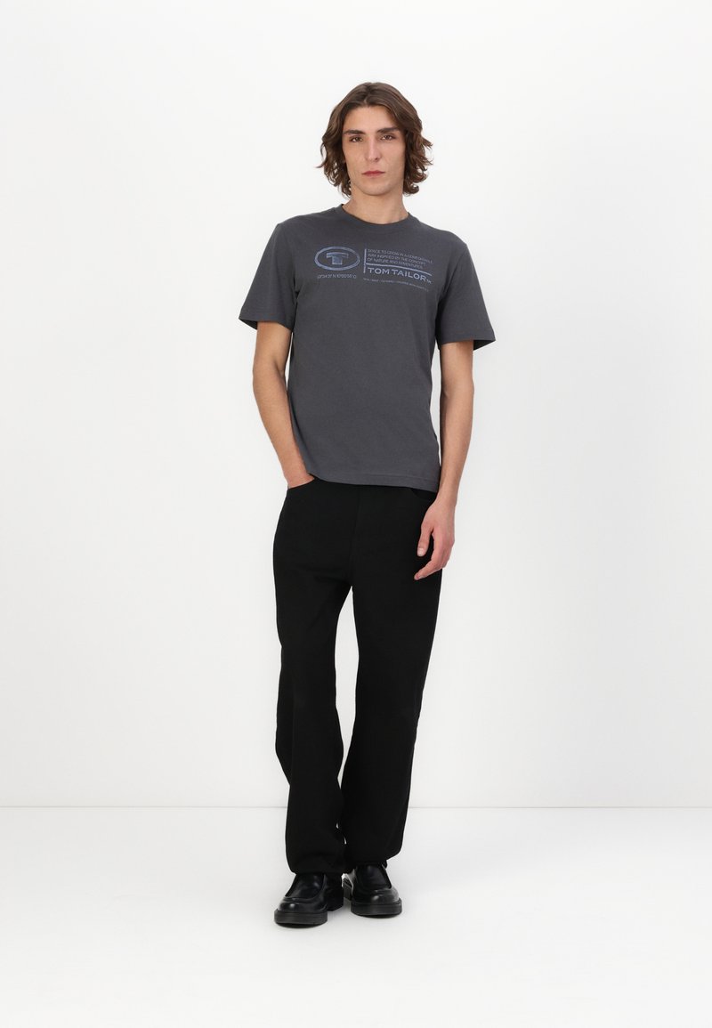 Camiseta de algodón gris con diseño de texto azul, combinada con pantalones anchos negros. El modelo está de pie en un fondo neutro, con las manos en los bolsillos.