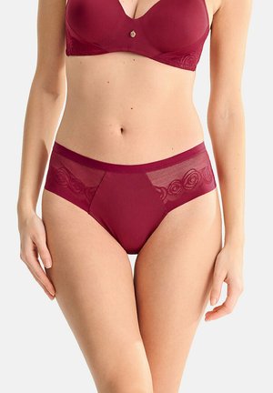 Femme portant un soutien-gorge bordeaux assorti et une culotte taille haute avec des détails en dentelle florale, debout sur un fond clair uni.