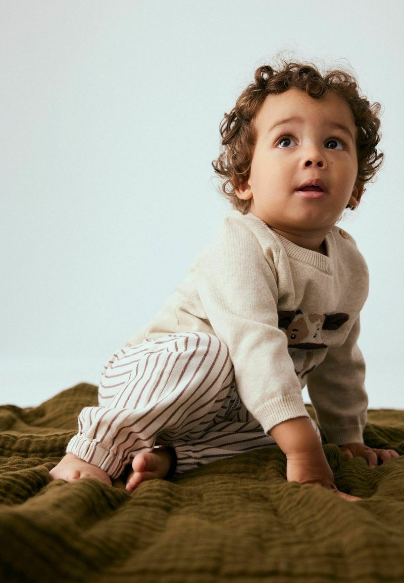 Tout-petit aux cheveux bouclés portant un pull beige avec un motif animalier et un pantalon rayé rampant sur une couverture texturée vert olive.