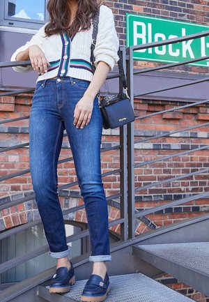 Femme portant un jean bleu retroussé, des mocassins bleu marine, un cardigan blanc avec des rayures colorées, et portant un petit sac noir en bandoulière dans les escaliers.