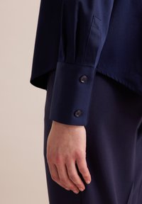 Marineblauwe shirt met een ontspannen pasvorm, voorzien van een puntige kraag en knoopsluitingen bij de mouwen. De stof lijkt glad met een subtiele glans.