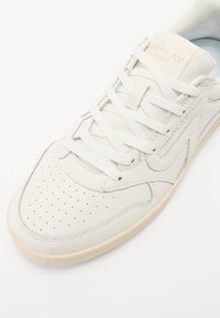 White leather sneaker with perforated toe, flat laces, light beige accents, padded collar, and embossed logo on the tongue.