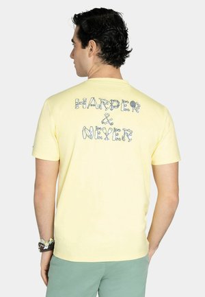 Man met donker krullend haar draagt een lichtgeel T-shirt met de tekst "HARDER & NEXER" gemaakt van geïllustreerd gereedschap op de rug.