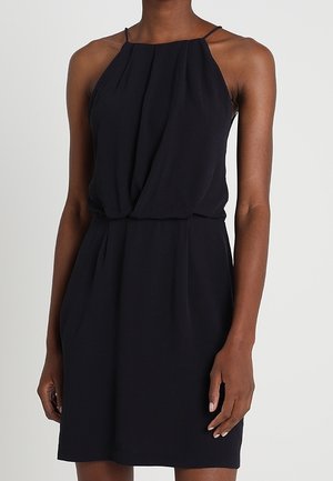 Femme portant une robe noire sans manches avec de fines bretelles et une taille froncée, debout devant un fond clair uni.