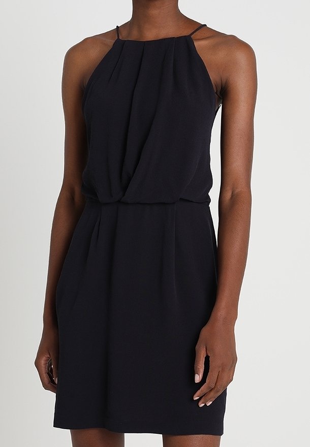 Femme portant une robe noire sans manches avec de fines bretelles et une taille froncée, debout devant un fond clair uni.