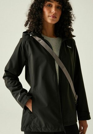 Woman wearing a black waterproof jacket over a gray shirt, with a gray crossbody bag strap, standing with one hand in pocket.