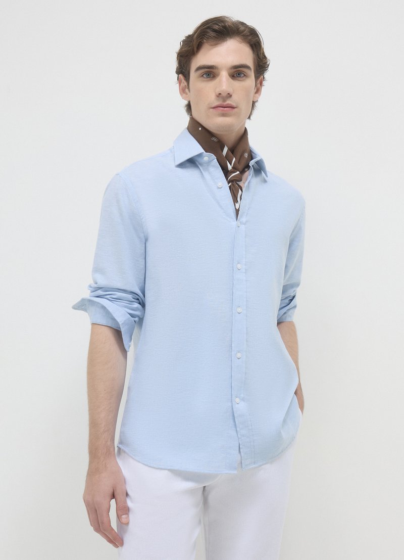 Chemise bleu clair à boutons avec manches retroussées, en tissu texturé ; présente une écharpe marron à motifs nouée autour du cou.